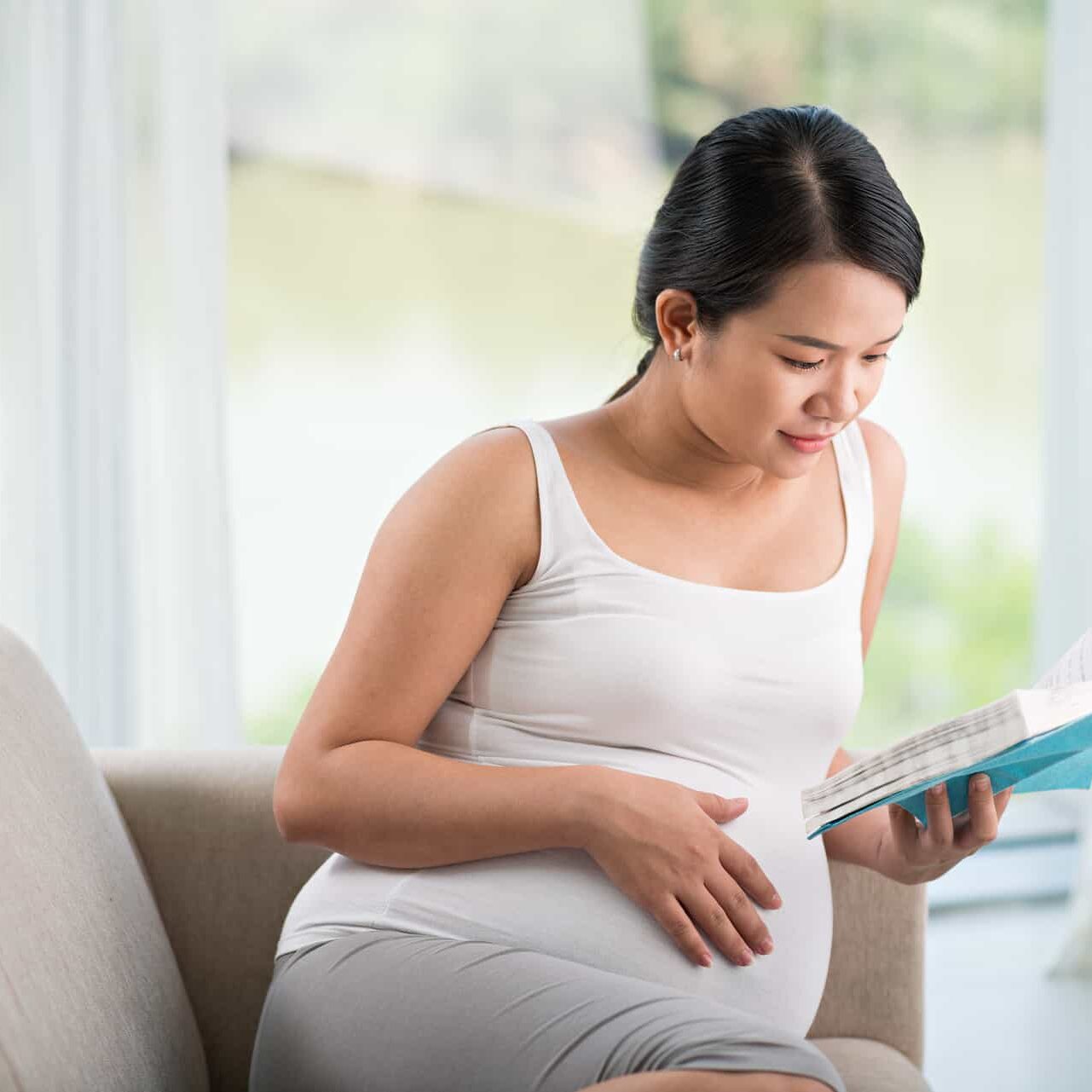 A pregnant woman sitting on a couch reading a book.